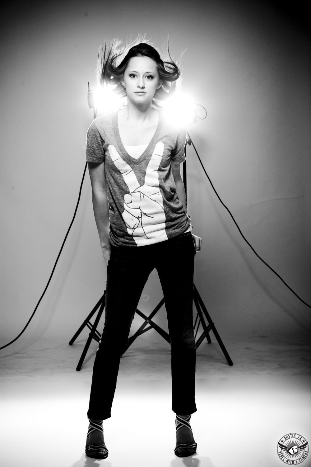 studio image of fashion model in black and white on white background with studio lights positioned behind model.  Model's hair is flipped up and is wearing a grey shirt with a hand  on the shirt  making a peace sign